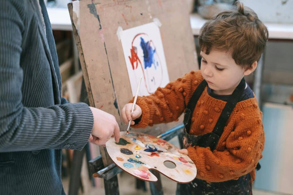 A little boy mixes paint on a wooden palette, with the help of an adult who points to the paint to be mixed.