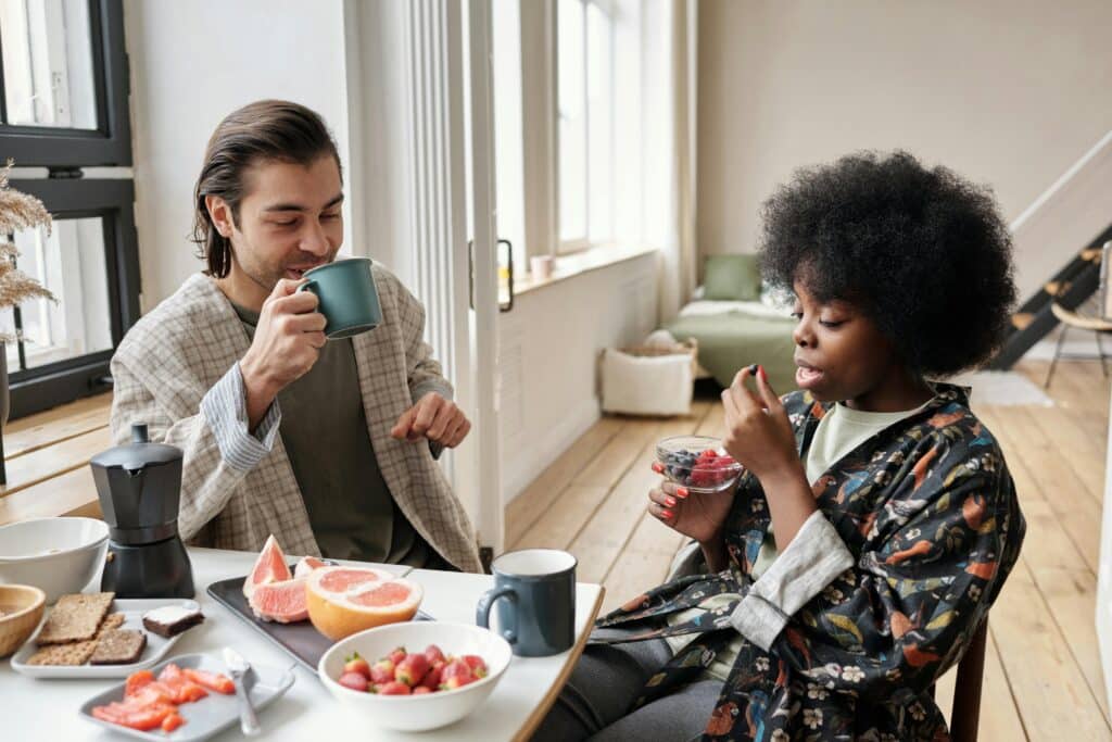 A young dark-haired man and a young black woman are having breakfast at the table. The man drinks a cup of coffee while the woman eats a bowl of red fruit. There is fruit on the table, including grapefruit and strawberries, as well as slices of bread. Their flat is bright, with white walls and light parquet flooring.