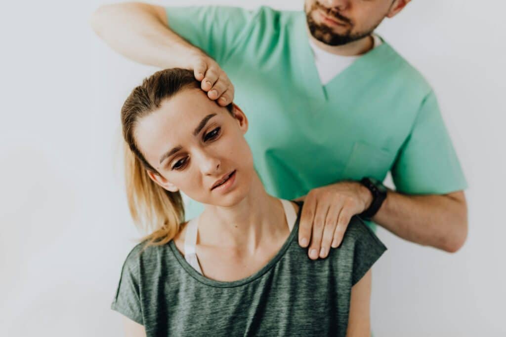 A young woman in a dark green T-shirt is being examined by a physiotherapist in a green lab coat. The practitioner holds her shoulder with one hand and the side of her head with the other. He stretches her neck.