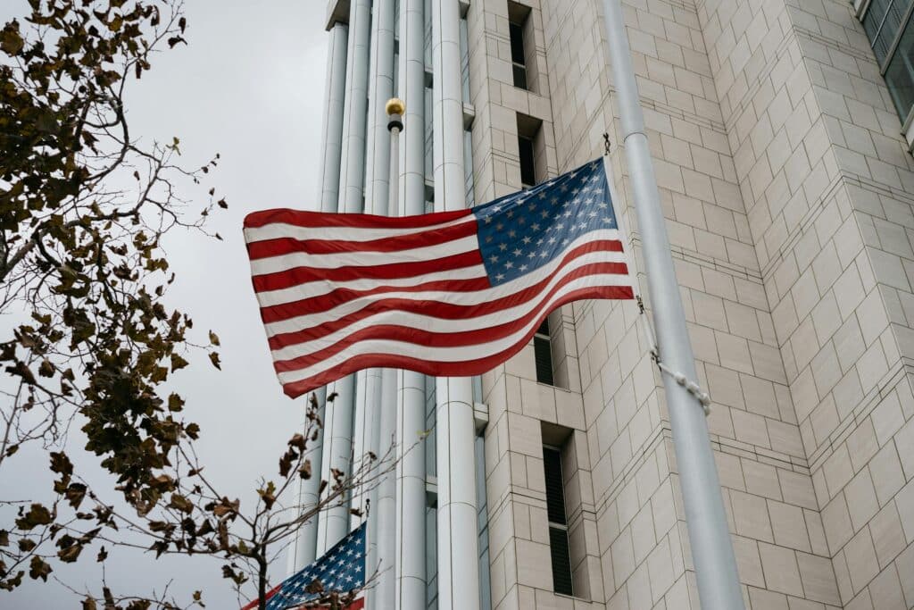 Photo of a raised American flag fluttering in the wind, taken in Anaheim in the United States, certainly in autumn. A tree is losing its leaves on the left of the image. The flag stands on a pole opposite a white concrete building.