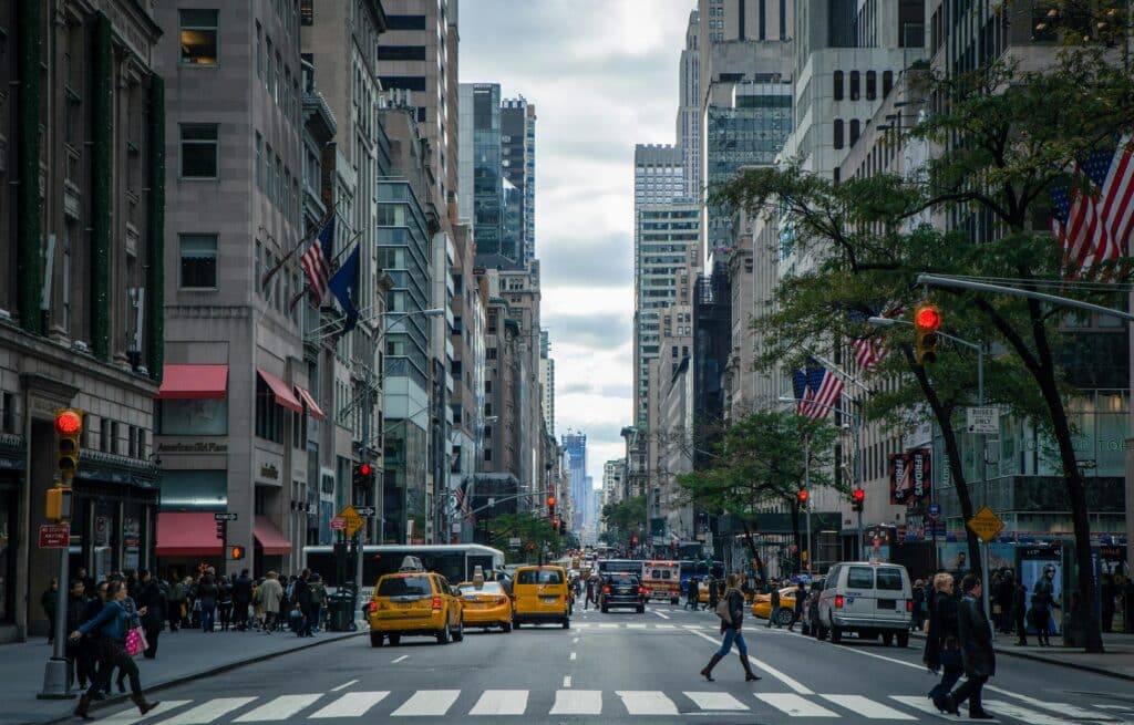 The photo was taken in the centre of a long, wide avenue in New York in the United States, with a pedestrian crossing in the foreground, yellow taxis driving along the road and many people walking on the pavements. American flags are flying from the buildings along the avenue. The sky is fairly cloudy.