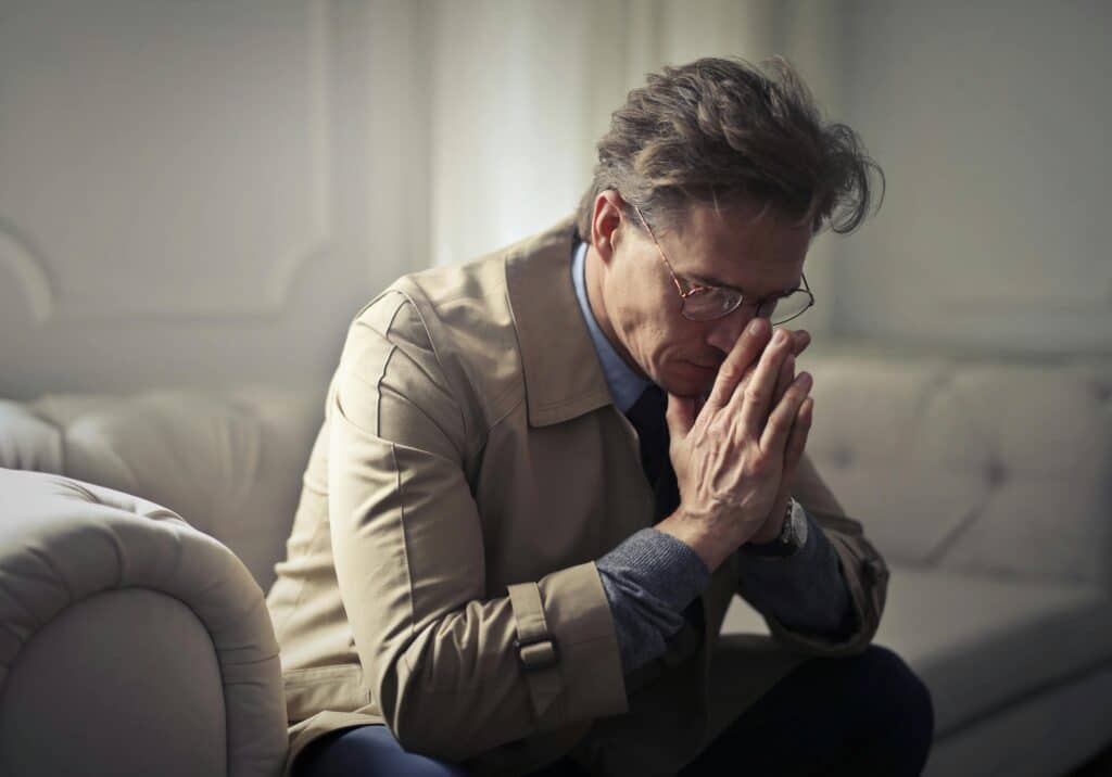 An elegant-looking man with greying hair is sitting on the sofa in his living room, supporting his face with his hands, looking thoughtful and worried.