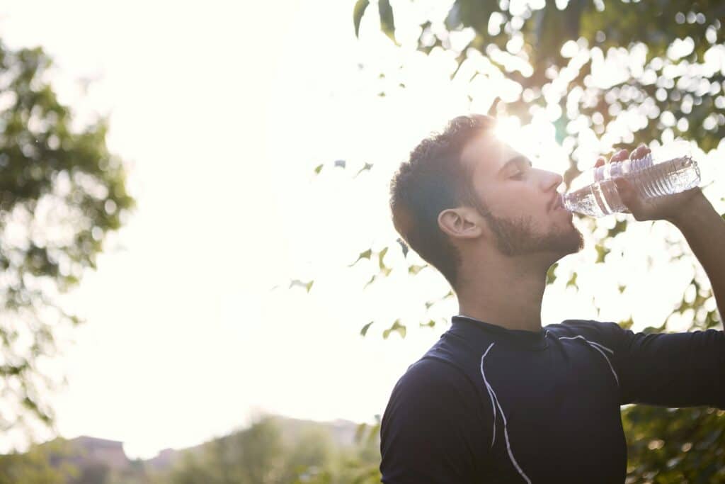 A dark-haired young man in a black sports outfit drinks from a bottle of water outside in the countryside.