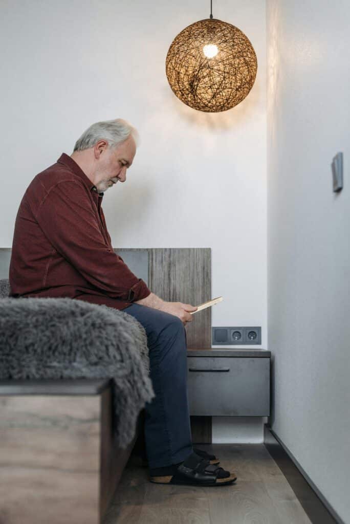 An old man is sitting on the edge of his bed, looking sad as he stares at a photograph in his hands. He is wearing a red shirt and blue jeans. He is looking at the photo by the light of a lamp hanging above him.