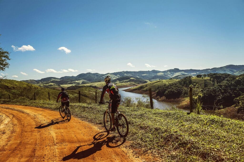 Two cyclists cycle along a red-sand country road, alongside a field near a lake.