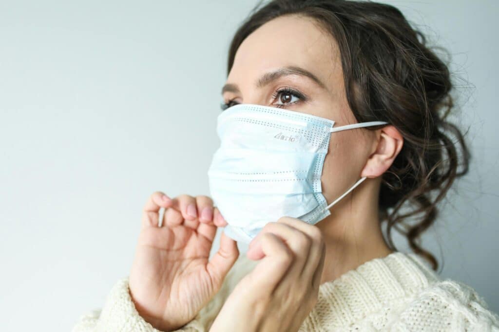 A dark-haired woman adjusts the surgical mask she is wearing. She is wearing a white knitted jumper and her hair is tied up in a low bun.