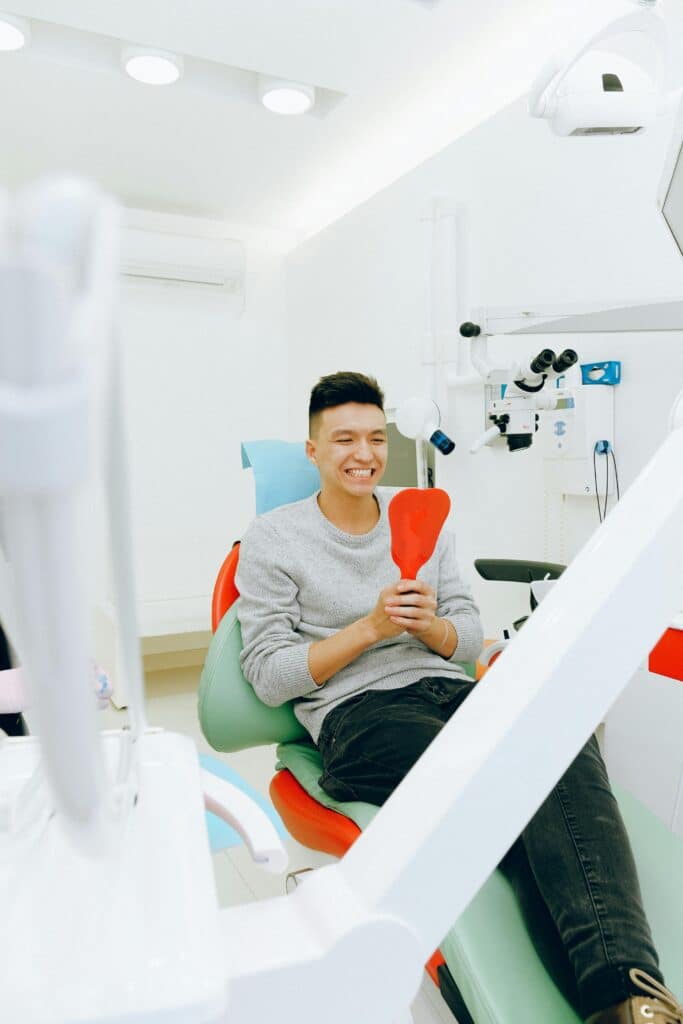 A patient sits in a dentist's chair and opens his mouth to look at his teeth in a mirror.