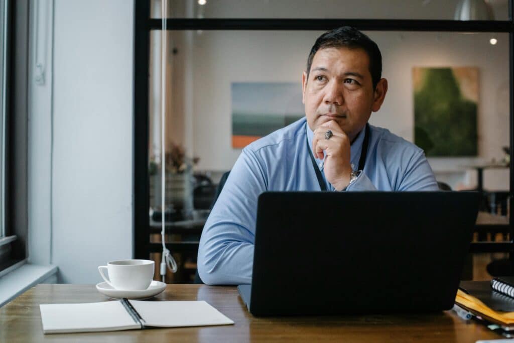 A man in a blue shirt sits at his desk and thinks as he works on his laptop. He has a cup of coffee and an open notebook beside him.