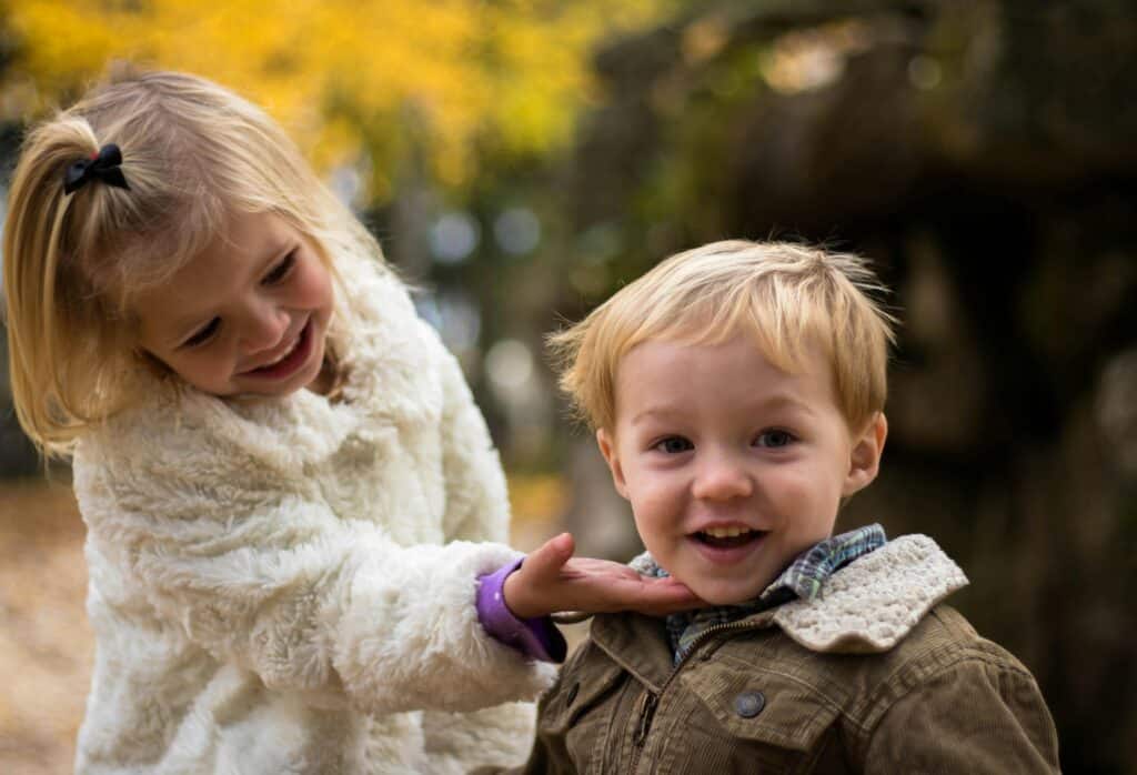 A little girl laughs with her little brother and holds him gently by the chin. They are dressed warmly for the autumn season.