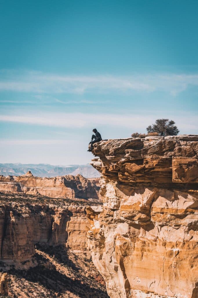 The photo was taken a little way off and shows a man sitting on the edge of a cliff with orange sand, staring into space. The sky is blue and the view is clear.