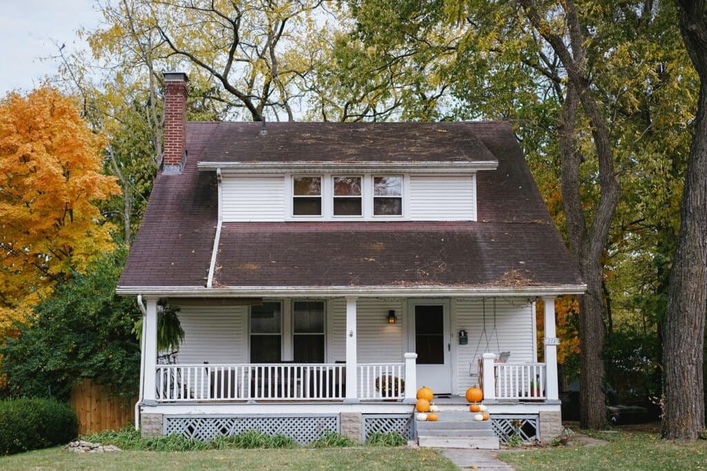 Front of a white house with a grey roof in typical American style. The house is surrounded by a garden and trees. Pumpkins line the entrance staircase.