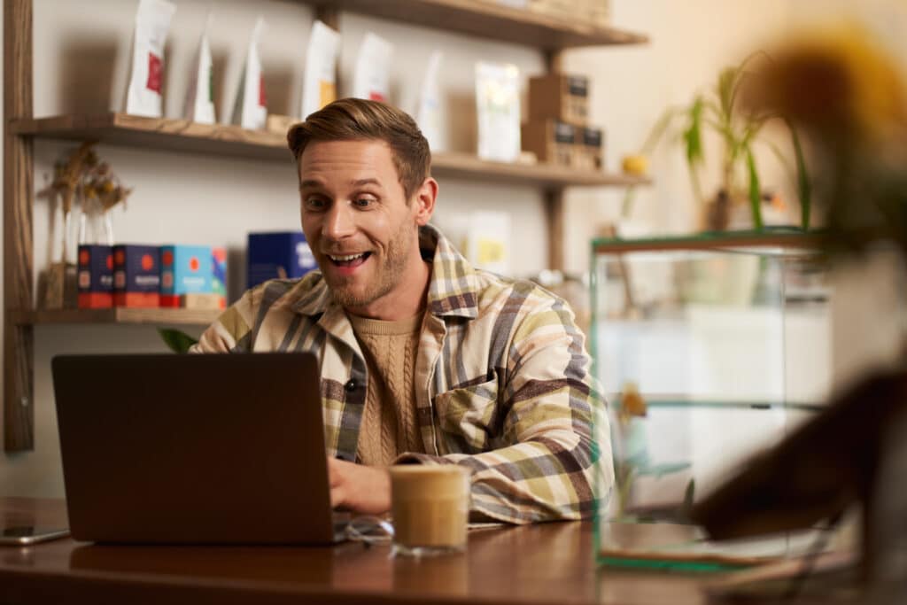 A blond-bearded man, a digital nomad, smiles as he types on his laptop, seated at a café table. He has a cup of coffee next to him and the shelves in the background display bags of different types of coffee.