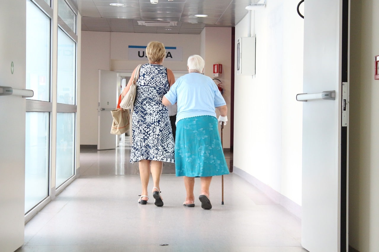 A woman in her sixties holds her elderly mother's arm and helps her walk down a hospital corridor to take her home.