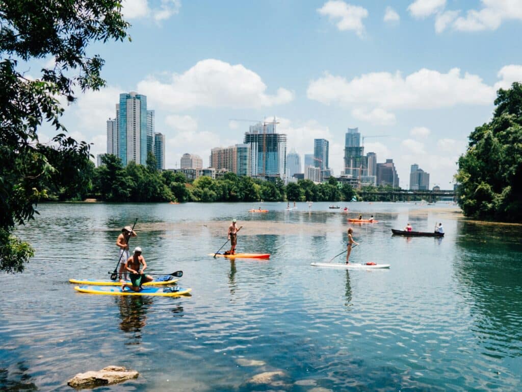 People paddleboarding on Lou Neff Point lake in Austin, USA. The lake is lined with trees. Tall buildings overlook the landscape. The weather is beautiful.