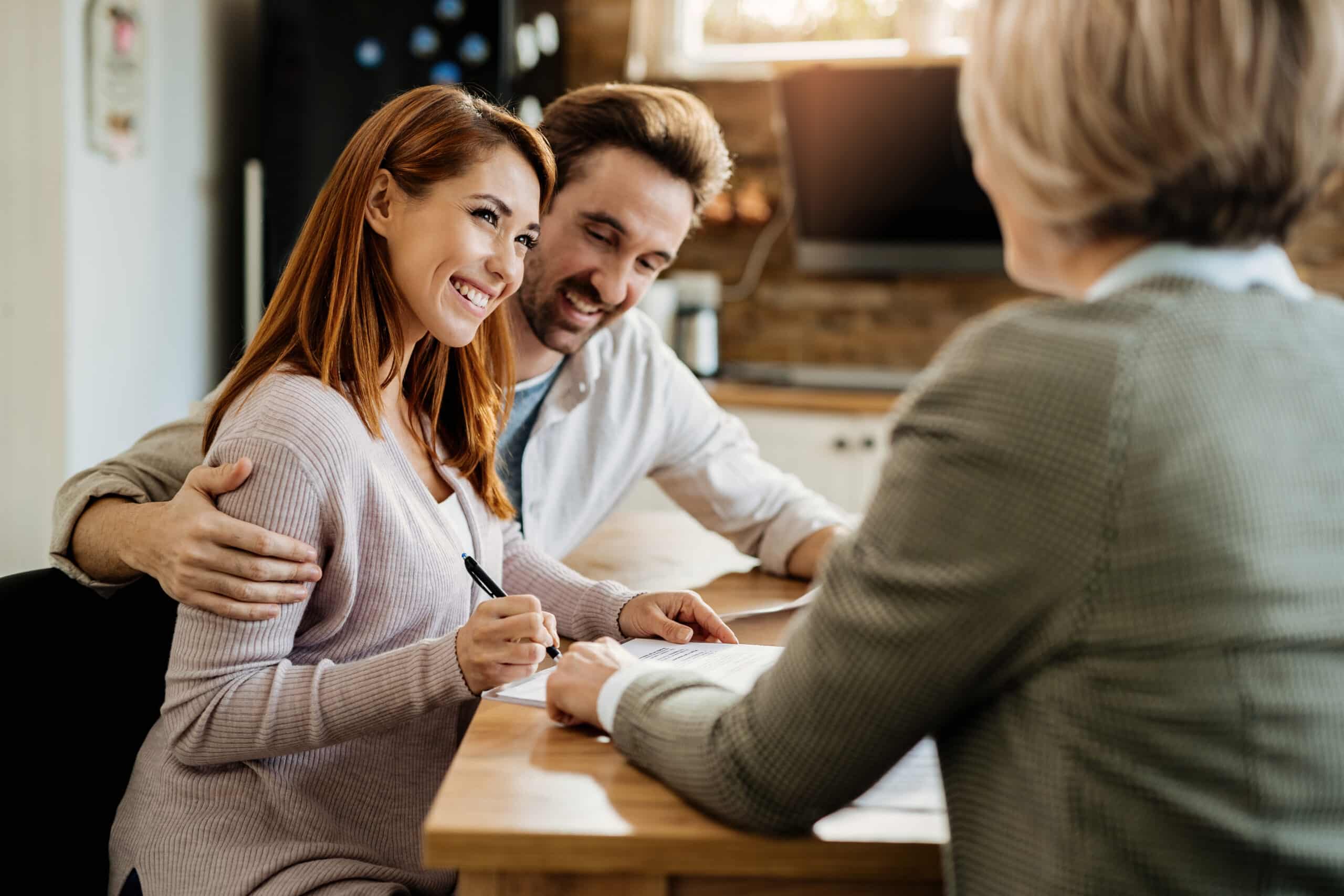 A happy young couple sign an insurance contract with an agent. The man wraps his arm around his wife's shoulder as he watches her sign the paper. The woman smiles at the agent.