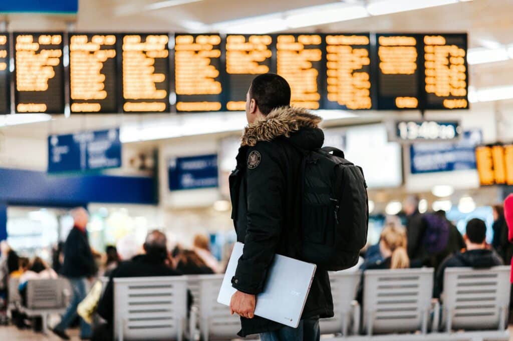 A young man wearing a parka and carrying a rucksack, holding a laptop computer in his hands, is standing in an airport and consulting the flight timetables displayed on the screen.