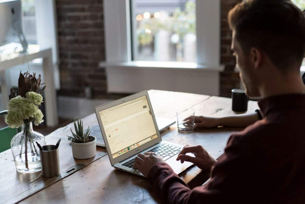 A red-haired man is sitting at a wooden table, typing notes on his laptop. We can't see his face, as the photo was taken over his shoulder. Another person is sitting next to him. Flowers and a plant are also on the table.