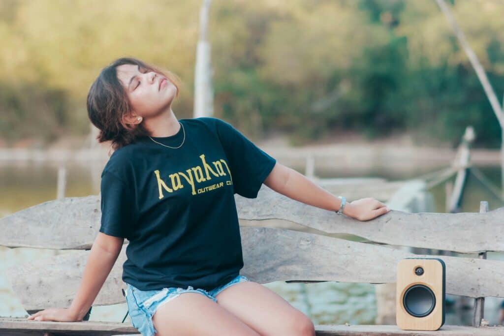 A young girl is absorbed in the music she is listening to through a loudspeaker. She is sitting on a wooden bench outside, leaning back with her eyes closed to better appreciate the music.