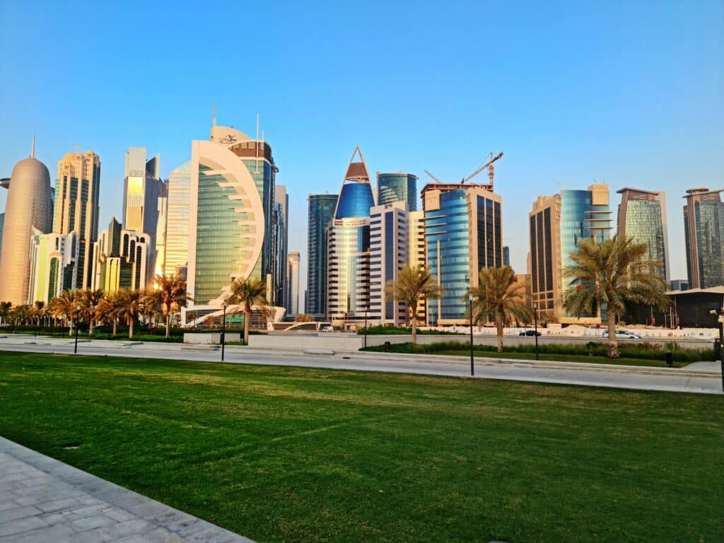 Skyscrapers of the city of Doha in Qatar, against a blue sky. The foreground shows a paved road with palm trees and a well-kept lawn.