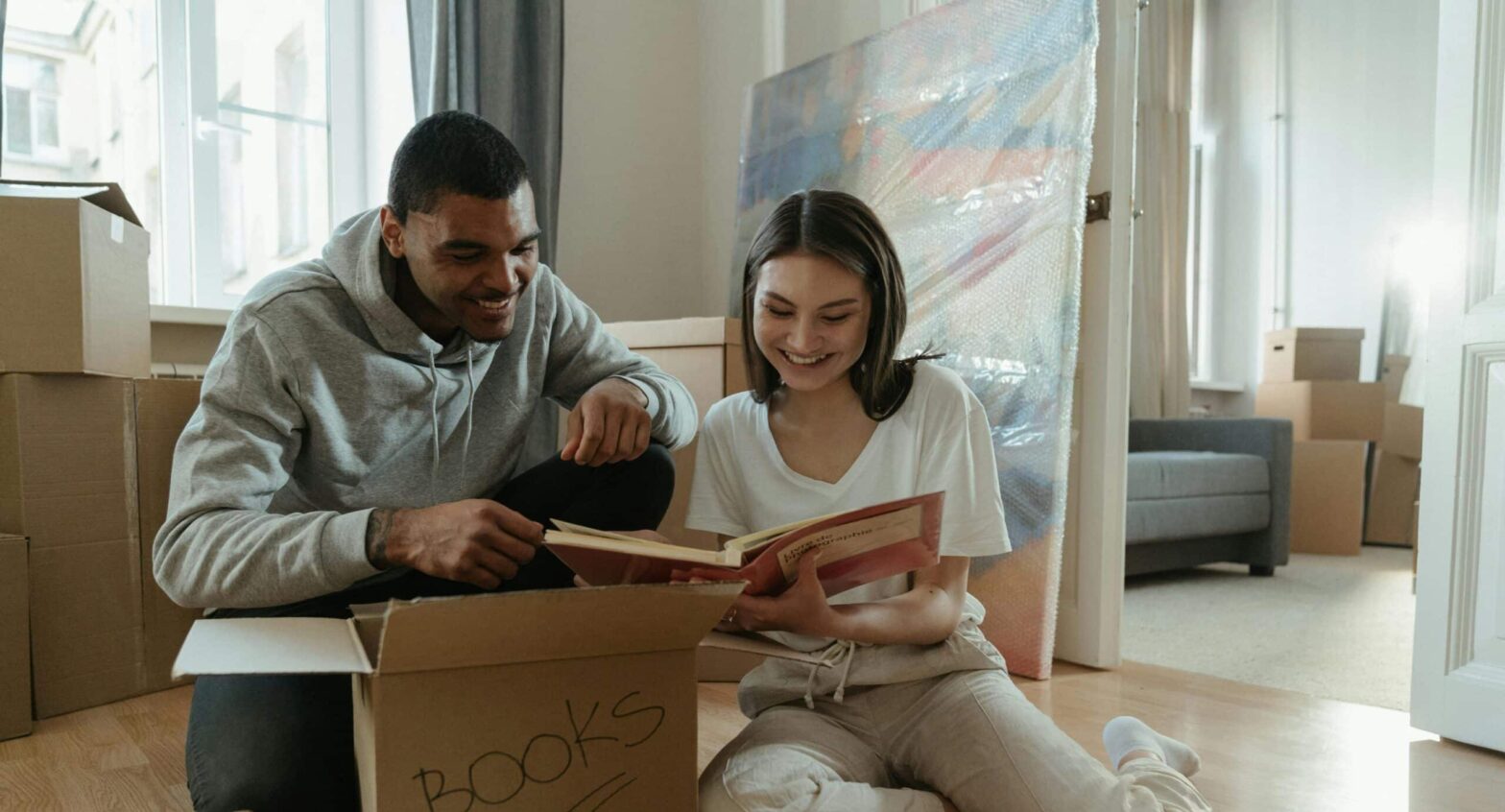 A young couple who have just moved into their new house are reminiscing as they read a book they have taken out of the boxes. They are sitting on the living room floor.