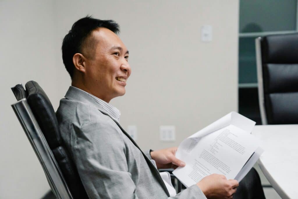 Asian man in a work suit smiles as he sits in his office chair.