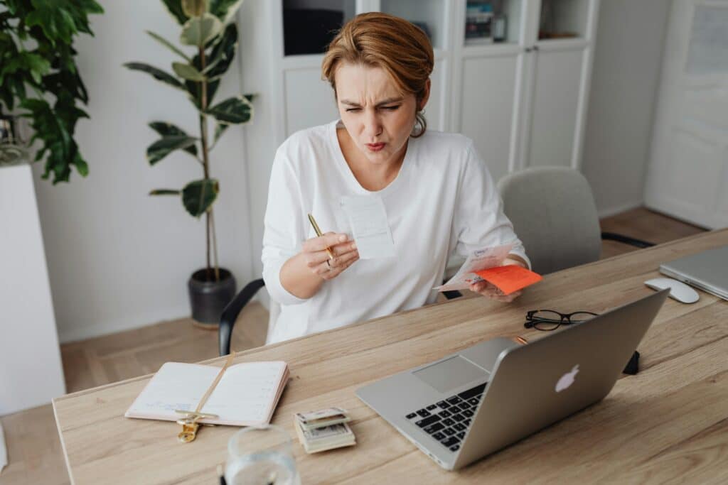 A woman sitting at her desk makes a face when she sees the amount written on an invoice she is holding in her hand. She is holding other credit card receipts in her other hand. On her desk are her computer, a notebook and a wad of banknotes.