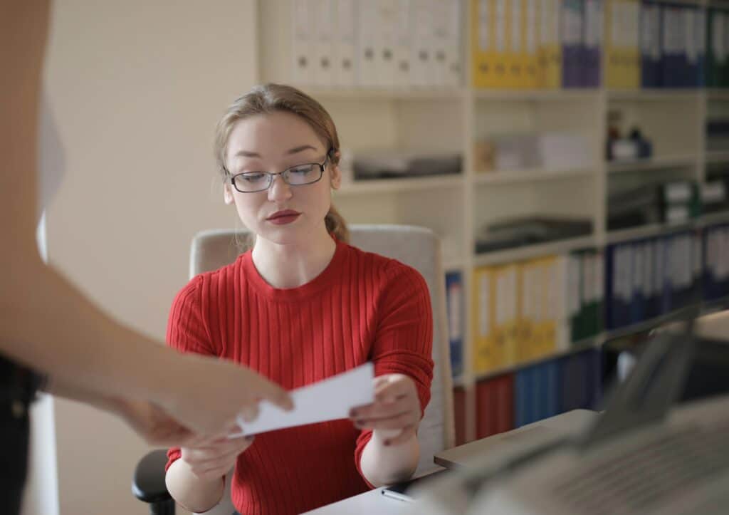 A young blonde woman wearing glasses is carefully reading a sheet of paper that has been handed to her. She is wearing a red jumper and matching lipstick. She is working on her laptop, with a bookcase full of binders and files behind her.