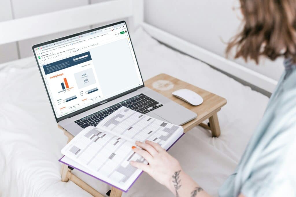 A woman with short hair and wearing a blue shirt is consulting her monthly budget on her laptop, sitting on her bed. We cannot see her face, as the photo was taken over her shoulder. She is consulting an organiser that she is holding in her hand.