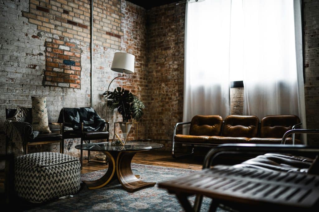 Vintage-style brick apartment. The furniture is a mix of old and modern styles, for example there is a leather armchair at the back of the room, the chairs are also leather and the coffee table is wooden. There is a simple white floor lamp and a Monstera plant as decoration.