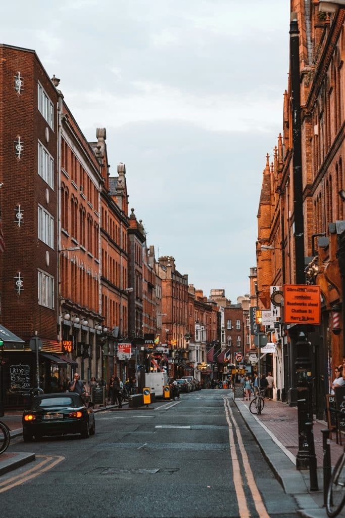 In this street in Dublin, Ireland, the buildings are medium-sized and built of orangey-red brick. A black car is parked at the side of the pavement. The street is not very busy and the sky is relatively grey.