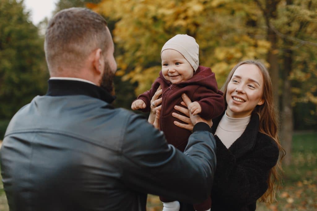 A young couple carries their babies with a smile, outside in the autumn season. They are wearing coats and are happy with the time they are spending together. The young woman has long blonde hair and blue eyes. The man has a beard and is dark brown.