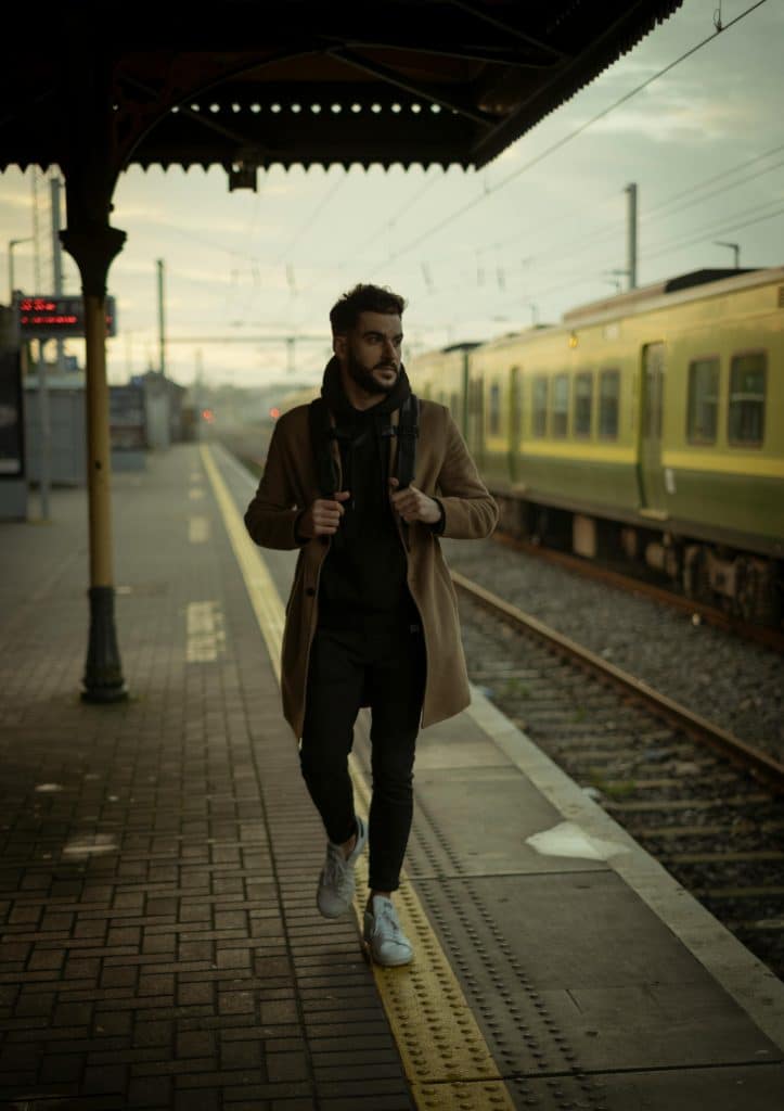 A bearded brown man wearing a rucksack walks along a train platform at a station in Dublin, Ireland. He is wearing a beige coat and black clothes. A yellow train is stopped on the other side of the track.