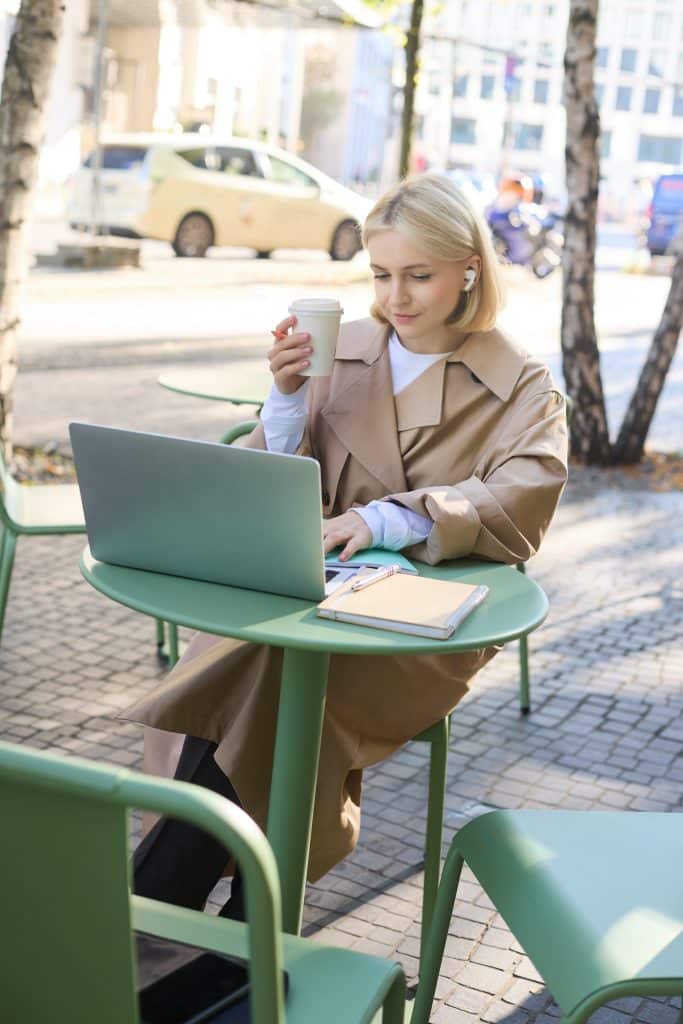 A young blonde woman wearing a beige trench coat is working outside on the terrace of a café. She is holding her drink in one hand and tapping on her laptop with the other. The outdoor furniture is sage green. The weather is clear and sunny.